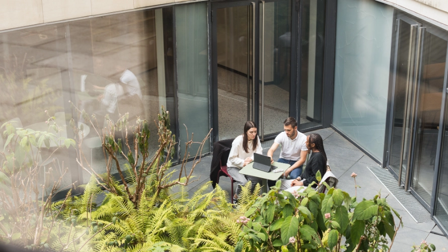 Two women, one man sit at a small outdoor table, looking at a laptop. Green foliage is in the foreground, large glass doors behind.