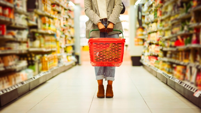 A shopper holding a red plastic shopping basket stands in the brightly lit aisle of a supermarket between two long, stocked shelves.